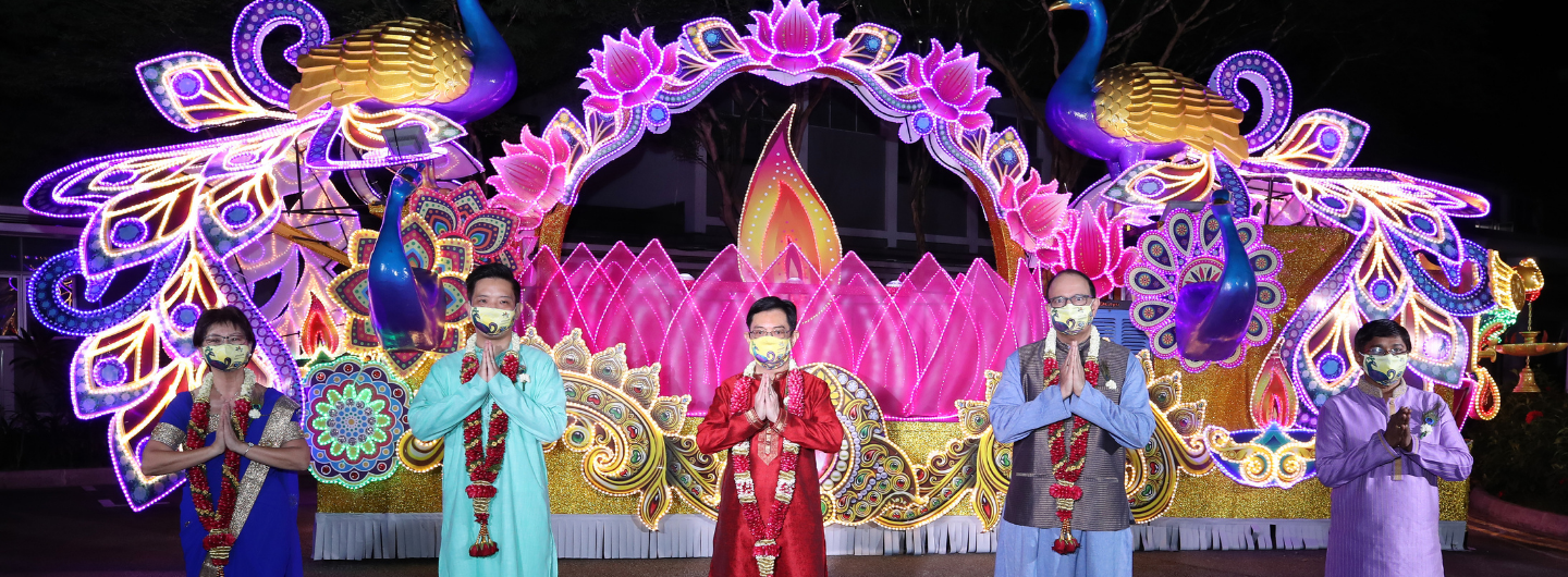 People with face masks wearing garlands, standing before illuminated peacock/lotus themed Diwali decorations.